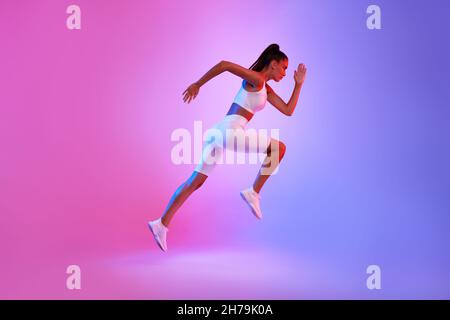 Determined Sporty Woman Running In Mid-Air Over Neon Background, Side-View Stock Photo