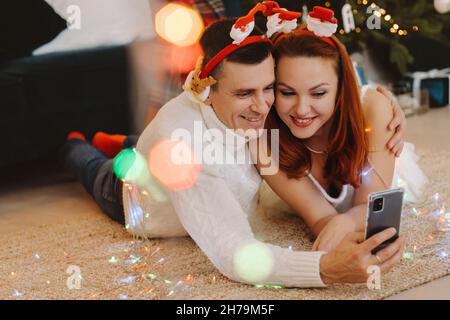 a happy married couple is lying on the floor at home near the Christmas tree and taking pictures of themselves. Stock Photo