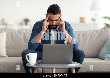 Young Arab man having headache, tired of working on laptop at home office Stock Photo