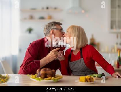 Affectionate senior man and his wife kissing and drinking wine while cooking festive meal for Christmas or Thanksgiving Stock Photo