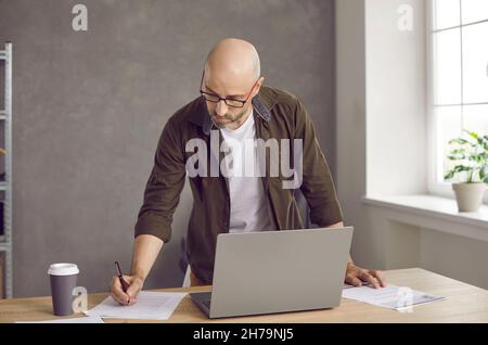 Serious focused businessman writes notes and uses laptop while working in his office. Stock Photo