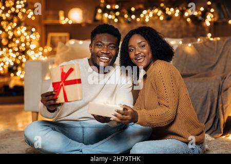 Cheerful young African American couple opening Christmas gift together in decorated living room Stock Photo