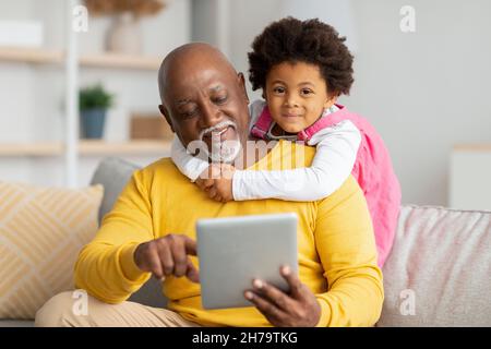 Happy african american preschooler girl hugs mature man surfing in internet or playing online game on tablet Stock Photo