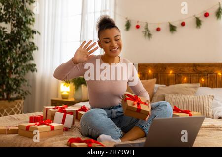 Positive african american lady offering gift box to laptop webcam, video calling on laptop, celebrating Christmas Stock Photo
