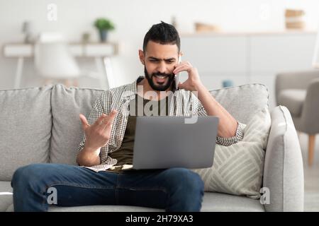 Handsome young Arab man calling on smartphone, using laptop for online meeting, working remotely from home Stock Photo