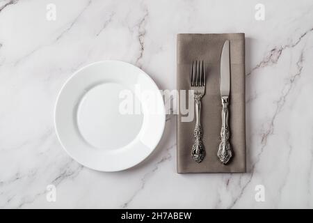 White porcelain plate near elegant vintage table knife and fork on a gray dinner napkin over marble tabletop. Set table. Empty clean ceramic plate. Stock Photo
