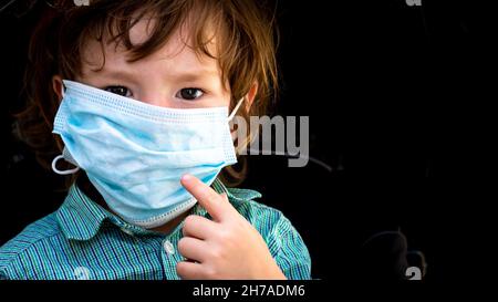 Toddler pointing to his face whilst wearing an adult size disposable face mask Stock Photo