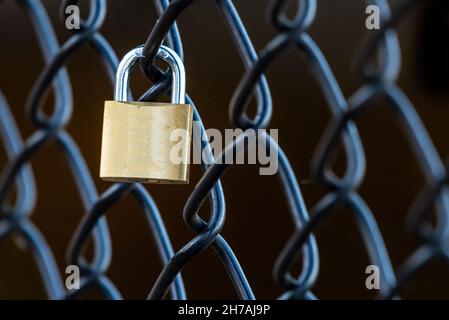 A brass padlock is attached to a chain-link fence. Stock Photo