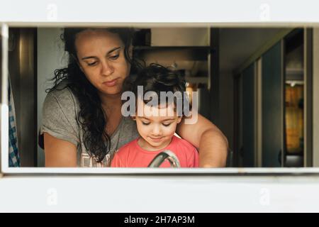 Loving mother teaching her little daughter to wash the dishes in the kitchen at home. Stock Photo