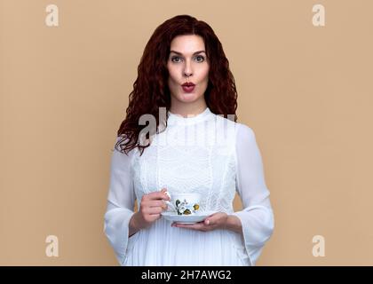 Woman with Fun Surprised Look Holding Vintage Teacup on Beige Background. Studio portrait of woman in romantic white dress holds vintage teacup. Stock Photo