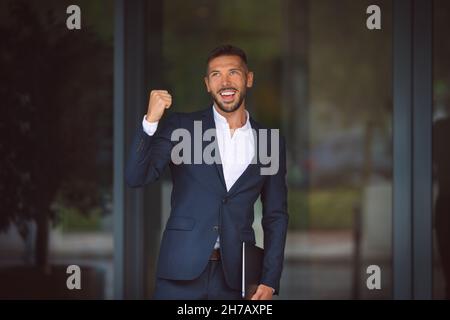 Happy businessman celebrating success and showing winner gesture. Young smiling office worker achieving goals. Leadership concept Stock Photo