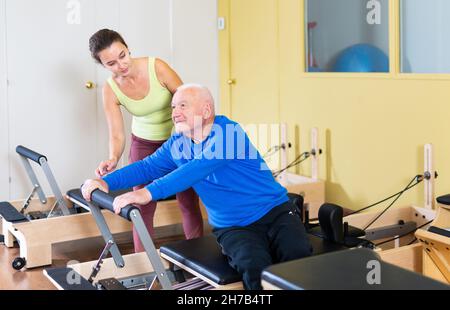 Woman pilates instructor helping aged man doing exercises on reformer Stock Photo