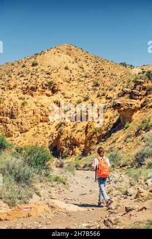 A woman with a backpack walks along a trail in a deserted canyon with red rocks. Hiking path and the dangers of solo trekking Stock Photo