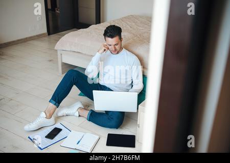 Caucasian man thoughtfully looking in laptop sitting on the floor in casual clothes. Workplace concept. Home concept. Distant education. Stock Photo