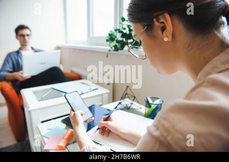 Couple working from home during a pandemic. Man standing at laptop and woman using phone makes notes. Standing face to face Stock Photo