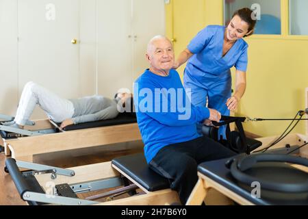 Woman physiatrist helping aged man doing exercises on reformer Stock Photo