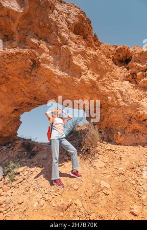 Tourist woman with backpack walks along a trail in a deserted canyon with majestic arch of red rocks. Hiking path and the dangers of solo trekking Stock Photo