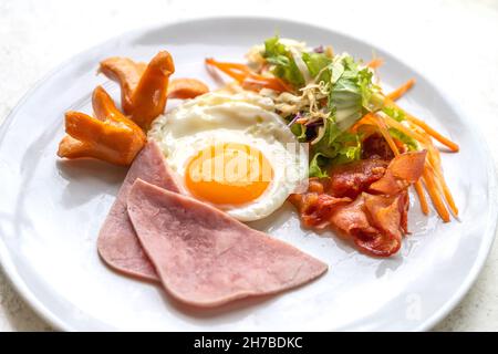 Breakfast menu, fried egg, ham, bacon, sausage, and salad. Breakfast on white plate on white stone table, natural light. Stock Photo