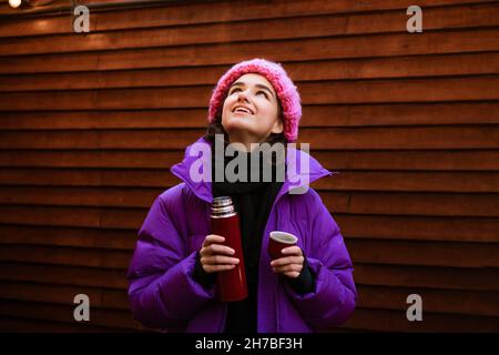 Young happy. A woman is drinking hot tea while holding a thermos on a winter day. Drinks to warm up in frosty weather in nature while walking around the city. In warm clothes and a knitted hat Stock Photo