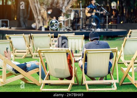 Back view of Asian young couple sit to relax on white deck chairs with tables for dinner in lawn is surrounded by shady green grass with blurred image Stock Photo