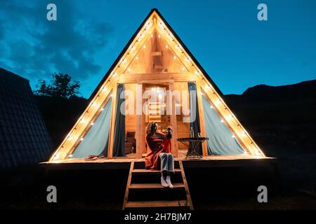 Woman drinking tea on the porch of a wooden lodge with lights of garlands in the evening. The concept of glamping and renting a chalet for weekend Stock Photo