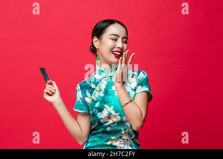 Young beautiful Asian woman wearing green oriental qipao dress holding credit card in red studio background for Chinese new year cashless shopping con Stock Photo