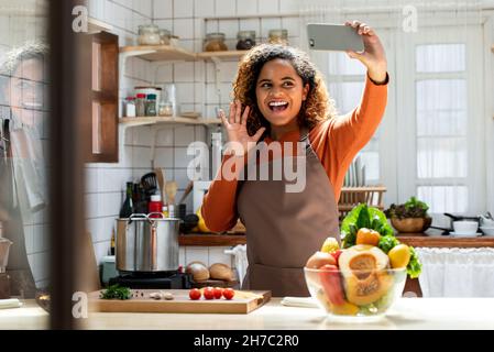 Young happy African American woman saying hi on video call while cooking in kitchen at home Stock Photo