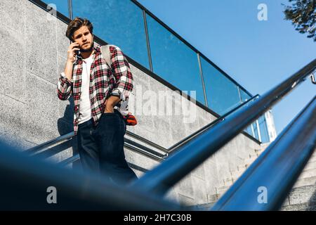Portrait of modern handsome young man speaking on phone. Hipster guy with mobile in his hand outdoors. Urban stairs, city street Stock Photo