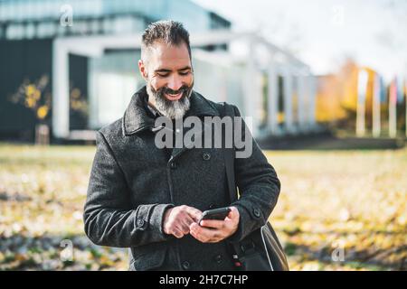 Businessman is using smartphone in front of company building in park. Happy businessman in autumn. Stock Photo