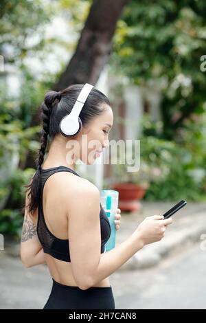 Pretty positive young woman in headphones turning on music on her smartphone to listen to during morning jog Stock Photo