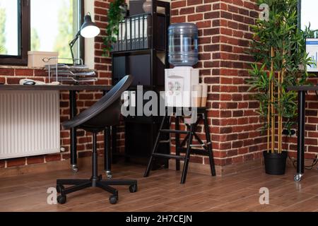 Close up of bookshelf and water dispenser in business office. Empty desk with decorations and watercooler in startup space, shelves with minimal decor. Decorative plants and refreshment. Stock Photo