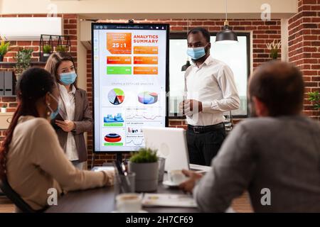 African american manager explaining management strategy working at business presentation in startup company office. Businessteam with medical face mask against covid19 brainstorming project ideas Stock Photo