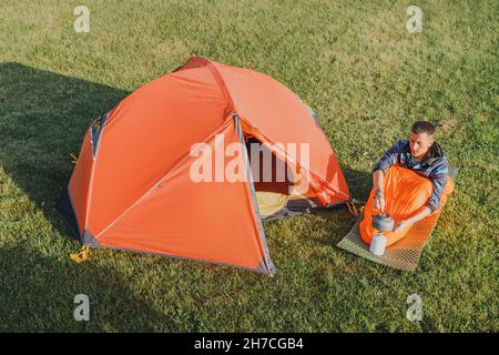 Aerial view of a man boiling tea on a cooking camping gas stove or burner, near open orange tent on a meadow at early morning Stock Photo