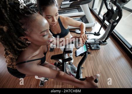 Coach installing application that helps to measure speed and distance on smartphone of client sitting on exercise bike Stock Photo