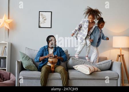 Portrait of two African-American girls jumping on sofa at home with happy father playing music, copy space Stock Photo