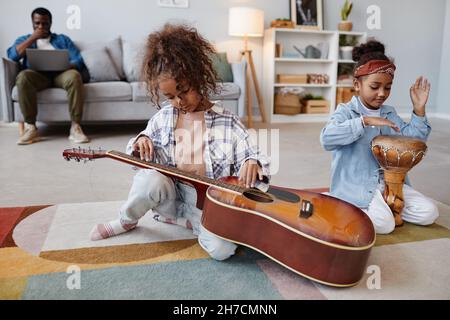 Full length portrait of two African-American girls playing with musical instruments on floor in cozy home interior Stock Photo
