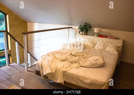 White unmade bed on large comfortable bed on the second floor of two-story country house in the woods. Modern interior of summer cottage with beveled Stock Photo