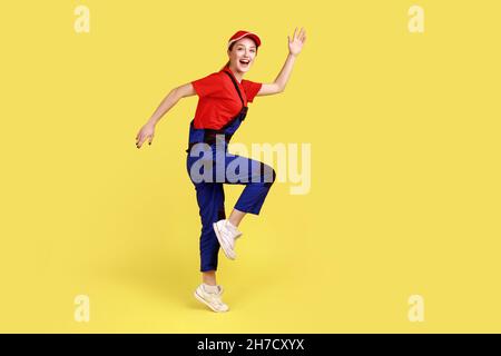 Side view portrait of excited worker woman standing on one leg with raised arms, looking at camera with happy expression, wearing overalls and red cap. Indoor studio shot isolated on yellow background Stock Photo