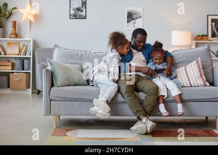 Full length portrait of happy African-American father relaxing on sofa with two cute daughters and using smartphone, copy space Stock Photo