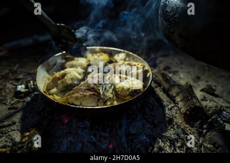 Large chunks of freshly caught fish are fried in a pan over a campfire, over coals, in the evening, at a camping. Lifestyle.  Stock Photo