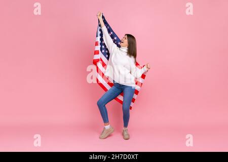 Full length photo of woman raised arms, holding american flag, celebrating national holiday, dancing, wearing white casual style sweater. Indoor studio shot isolated on pink background. Stock Photo
