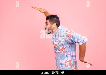 Side view of cheerful bearded man in blue casual shirt dancing, having fun and doing funny egyptian arm dance move, celebrating, rejoicing. Indoor studio shot isolated on pink background. Stock Photo