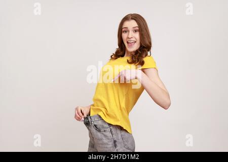 Profile portrait of beautiful young woman have willpower to loose weight, showing slim waist and pointing finger on big trousers, diet conception. Indoor studio shot isolated on gray background. Stock Photo