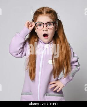 Portrait of surprised, amazed kid girl in pink jumpsuit with hood standing with mouth open holding her glasses with hand Stock Photo