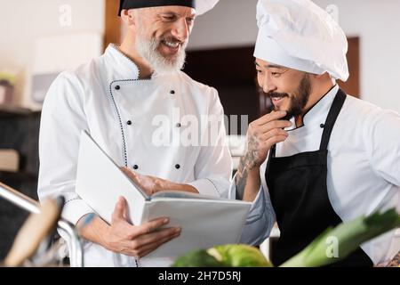 Cheerful interracial chefs working with cookbook in kitchen Stock Photo