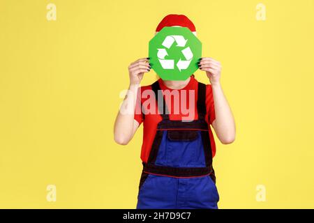 Portrait of unknown anonymous worker woman standing hiding her face behind green recycling sign, ecology, wearing overalls and red cap. Indoor studio shot isolated on yellow background. Stock Photo