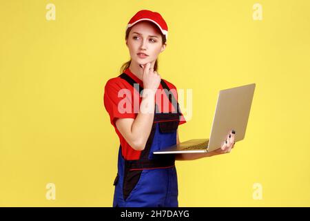 Full length thoughtful uncertain worker woman holding laptop, pondering decision, online order for repair services, wearing overalls and red cap. Indoor studio shot isolated on yellow background. Stock Photo