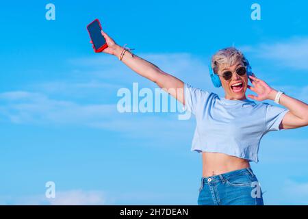 young blonde woman with short hair dressed in short t-shirt showing her belly button has fun and laughs with open arms while wearing wireless headphon Stock Photo