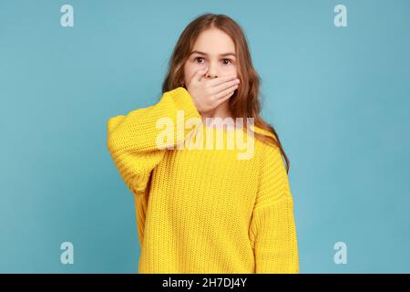 Portrait of little girl covering mouth with hand, afraid to say secret, child terrified to speak, wearing yellow casual style sweater. Indoor studio shot isolated on blue background. Stock Photo