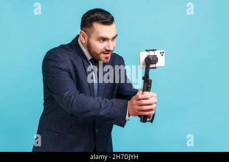 Young adult man blogger with beard wearing official style suit broadcasting livestream via smart phone and stedicam, talking to his followers. Indoor studio shot isolated on blue background. Stock Photo